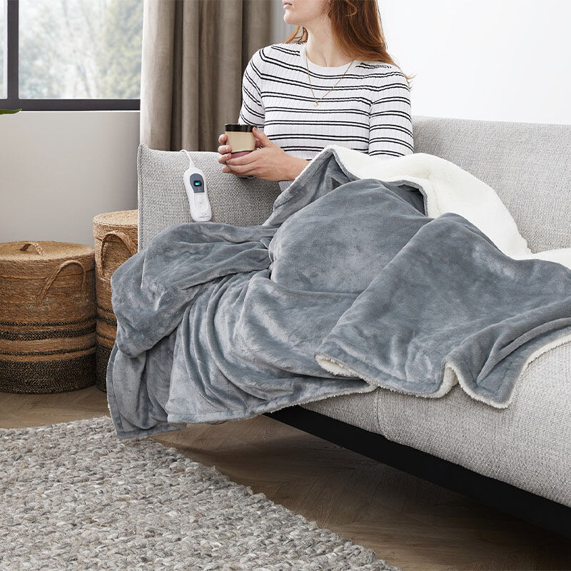 Een vrouw zit onder het SleepMed Elektrische Knuffeldeken in de lichtgrijs. 
